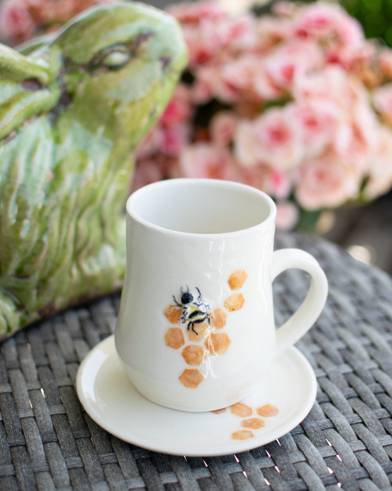 White mug with a bee design on a saucer, placed on a textured surface with flowers in the background.