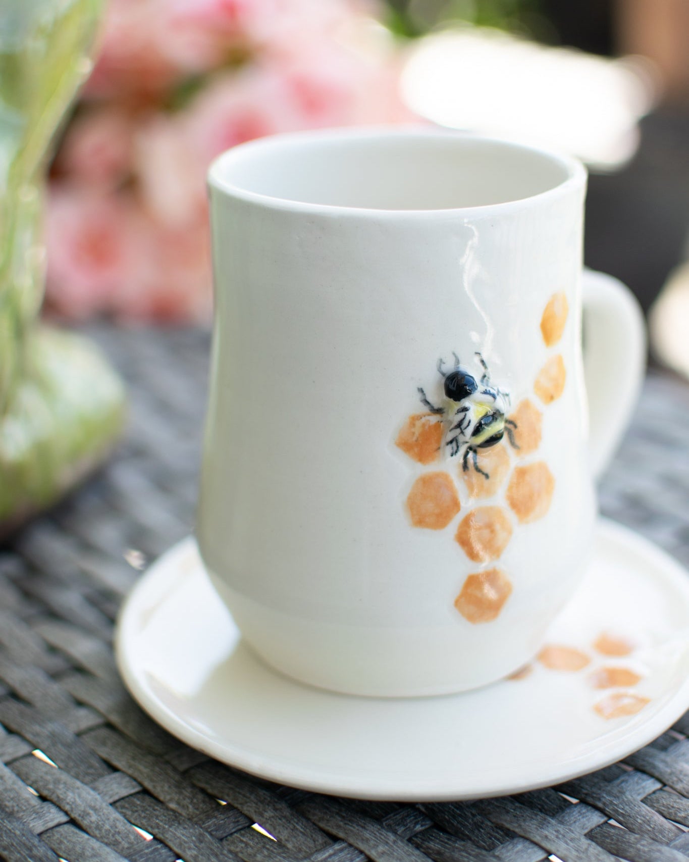 White mug with a bee design on a saucer, placed on a textured surface with flowers in the background.
