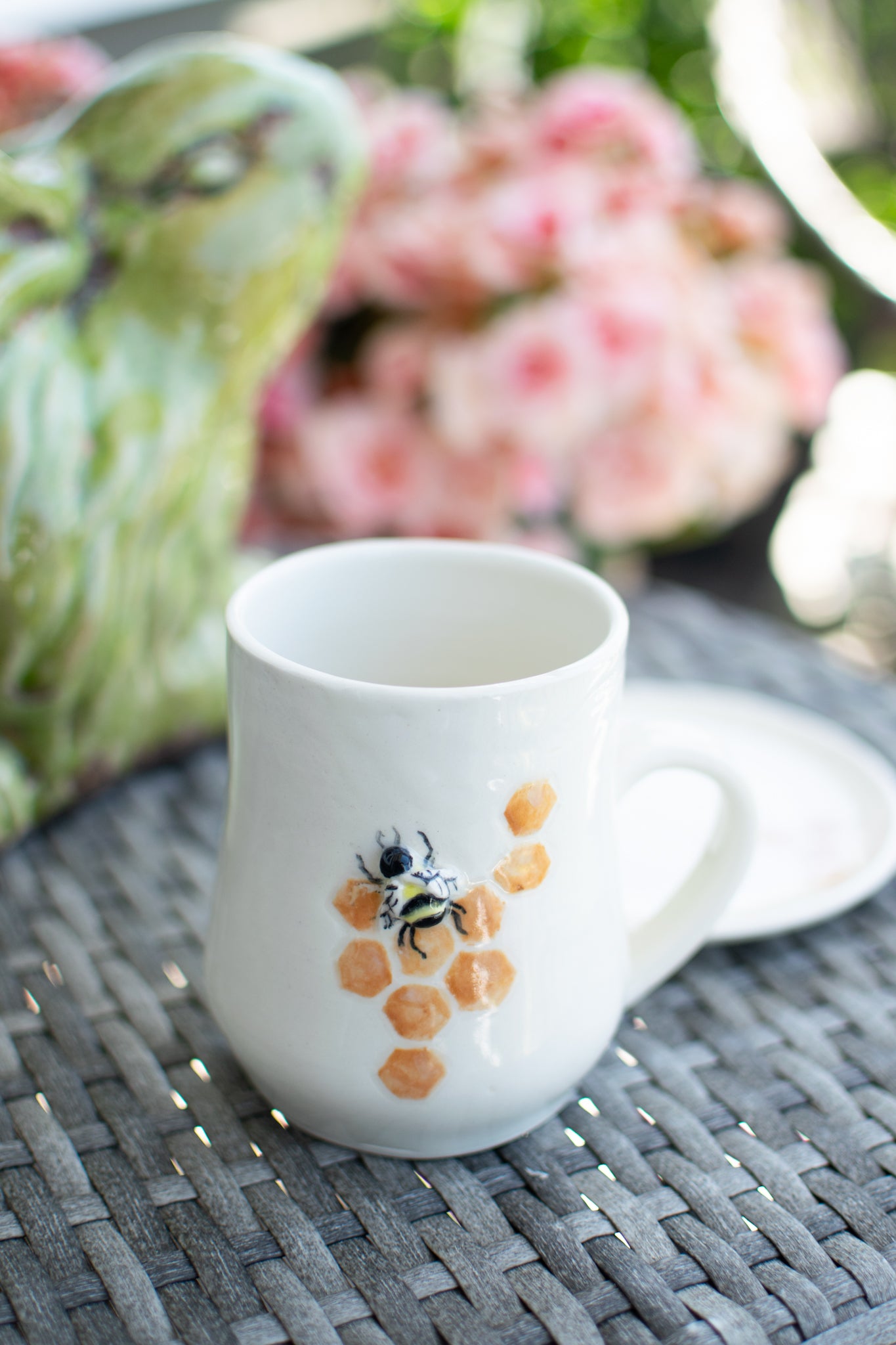 White mug with a bee design on a woven surface with flowers in the background