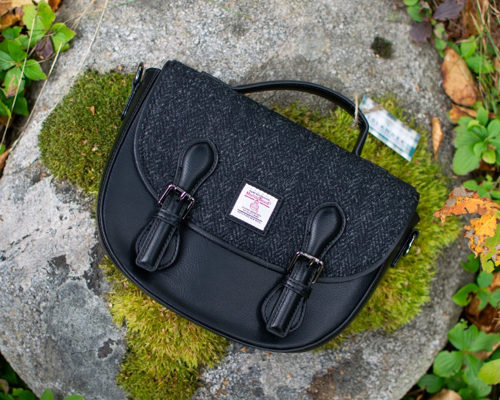 Black textured handbag on a stone surface with green moss and leaves.