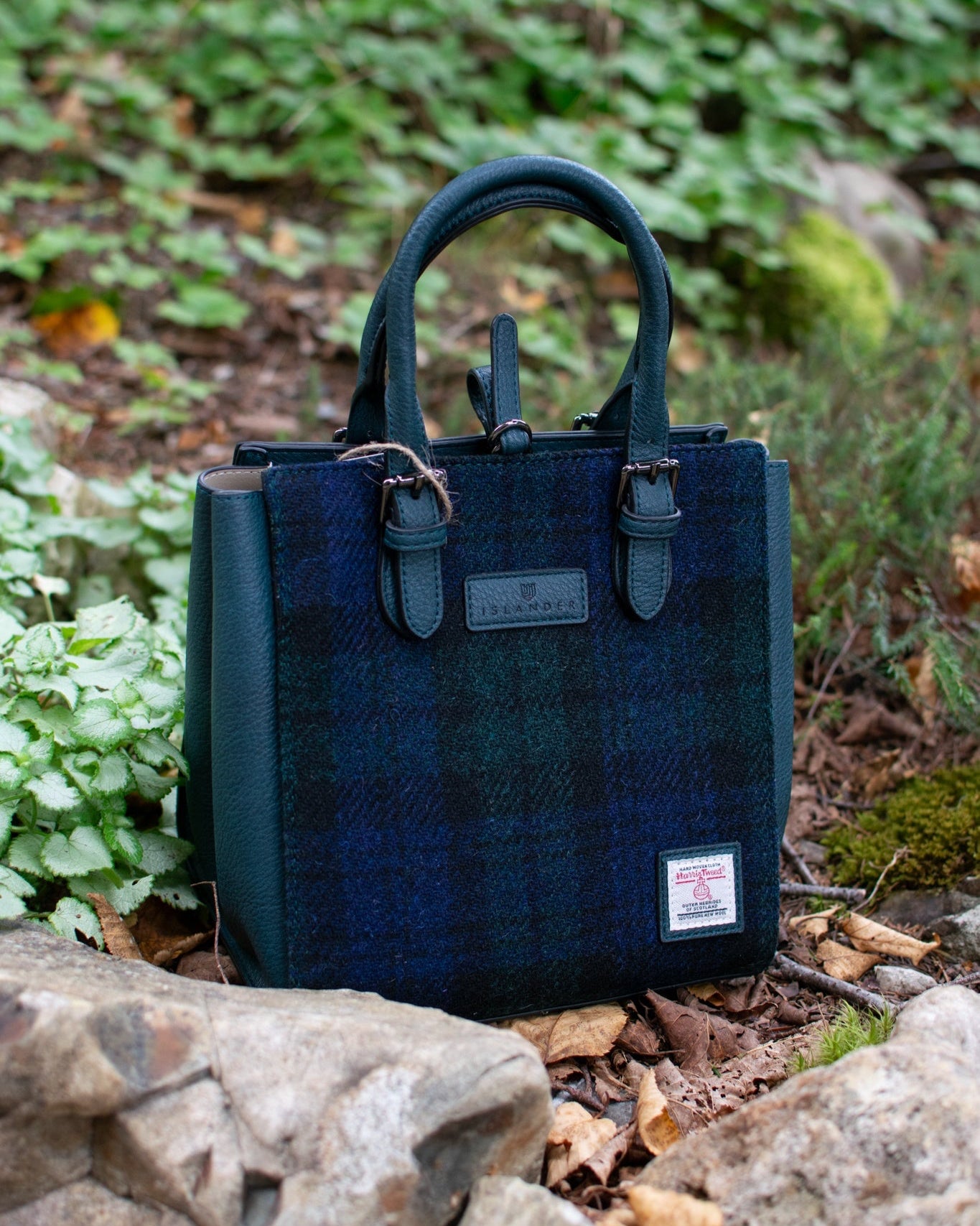 Blue plaid handbag with a visible brand label on a natural background of rocks and plants.