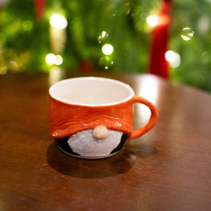 Gnome-shaped cup with a red handle on a wooden surface with a blurred Christmas tree in the background.
