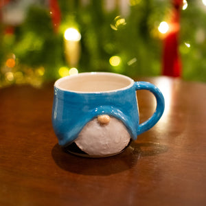 Blue gnome-shaped mug on a wooden surface with a blurred Christmas tree in the background.